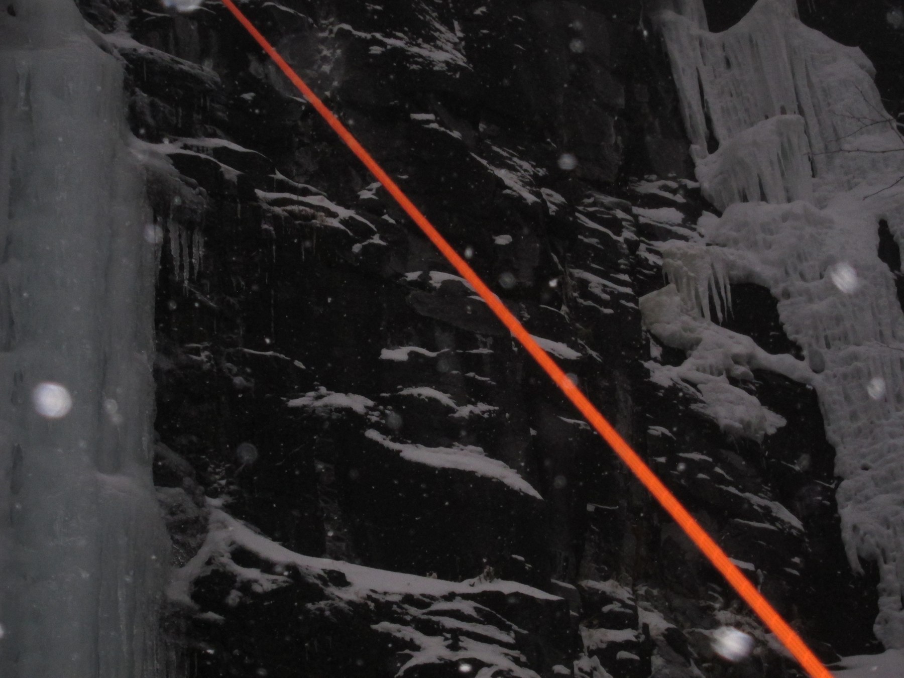 Orange climbing rope cutting diagonally across frozen winter rock face and icefalls in snowfall